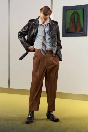 Model wearing black leather jacket, blue button-down shirt, striped tie, and brown trousers standing indoors.