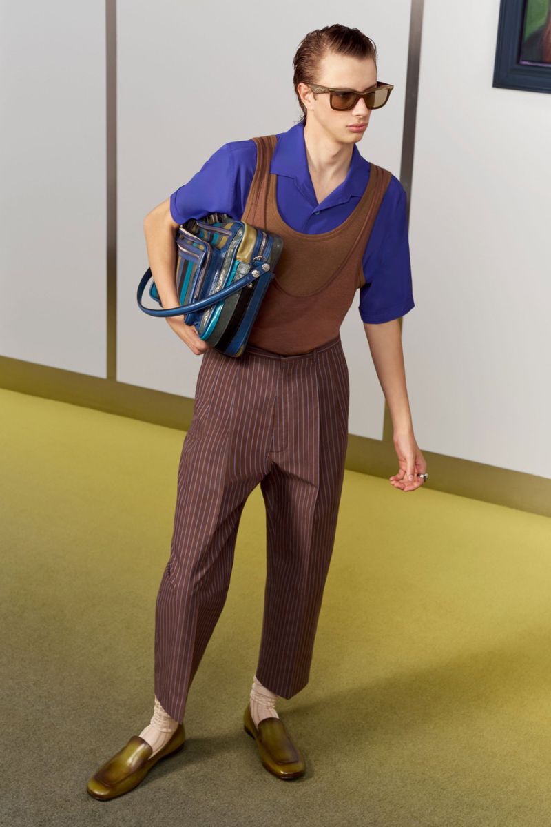 Model Pavlo Stinyk wearing a blue shirt, brown vest, and pinstripe trousers carrying a bag for Acne Studios.