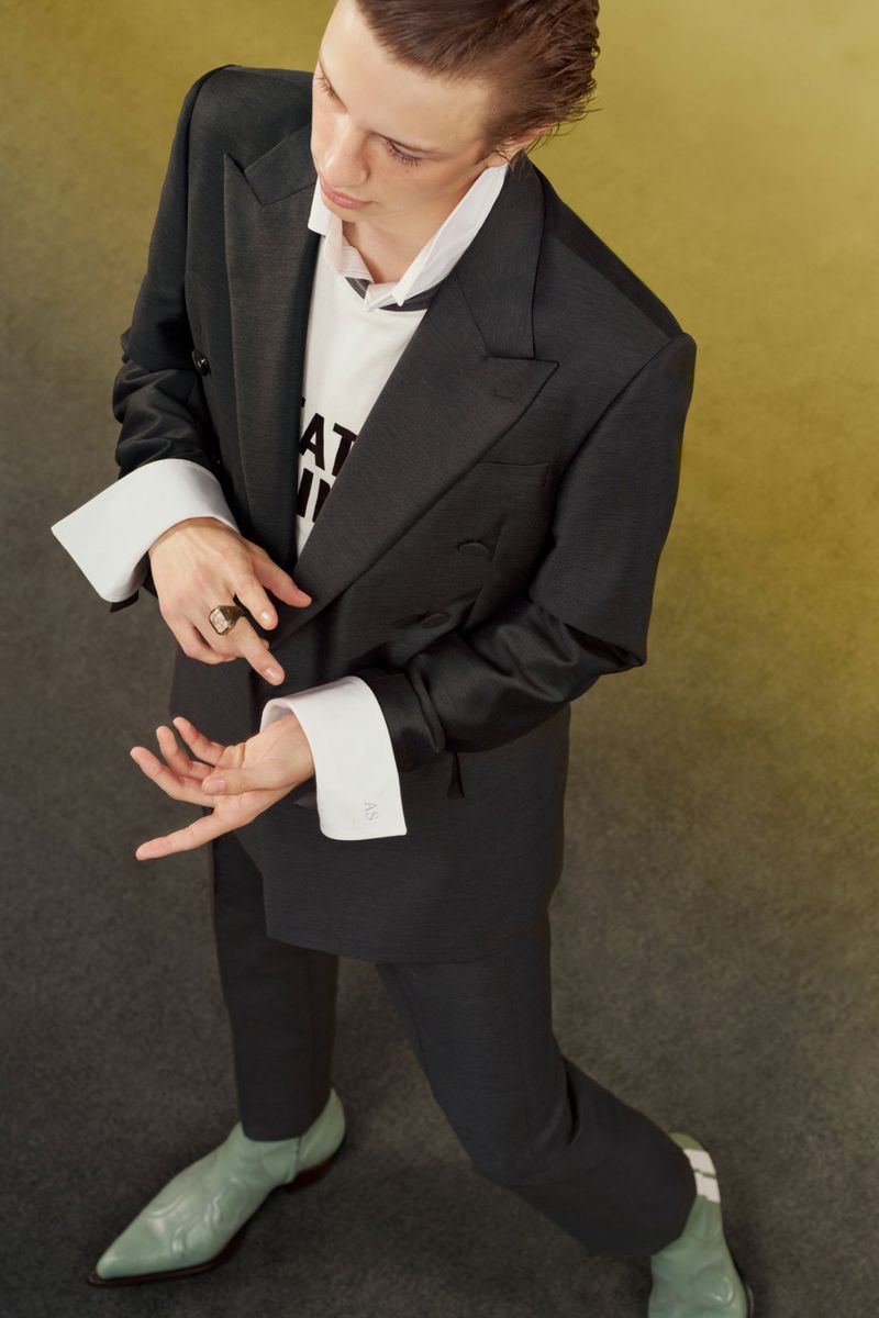 High-angle view of a male model wearing an oversized black suit with exaggerated white cuffs and pale green pointed boots.