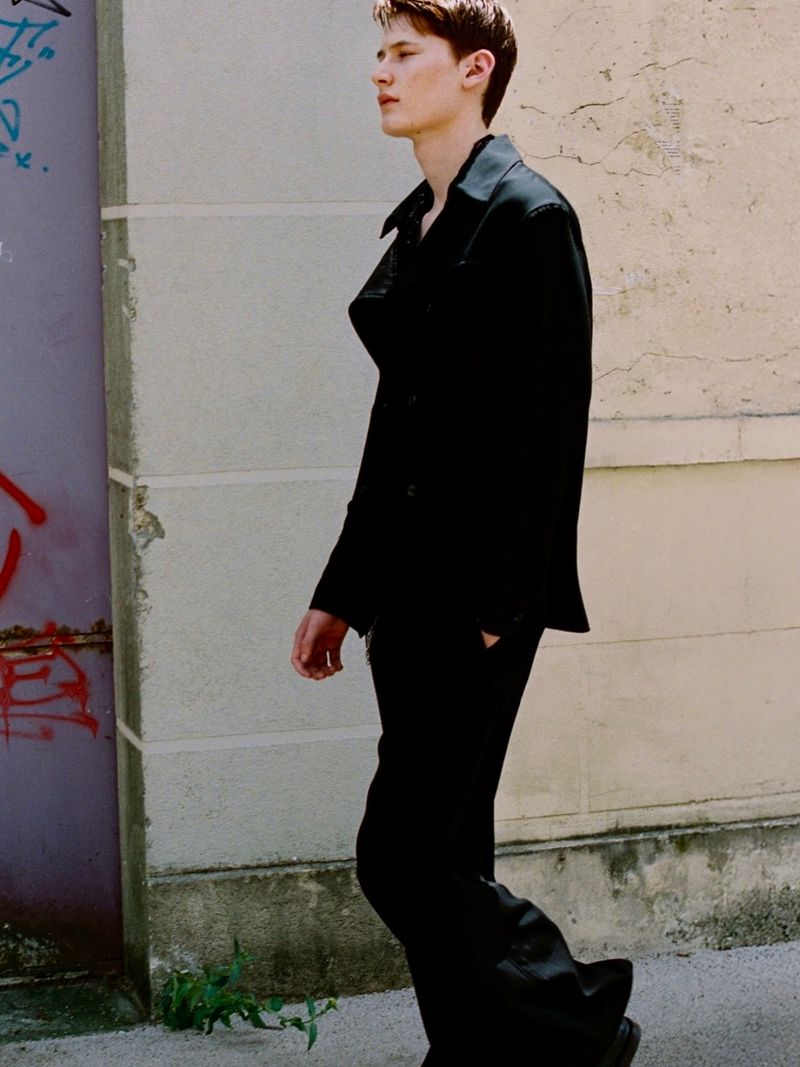 Young man in an all-black jacket and wide-leg trousers walking in profile alongside an exterior stucco wall with graffiti.