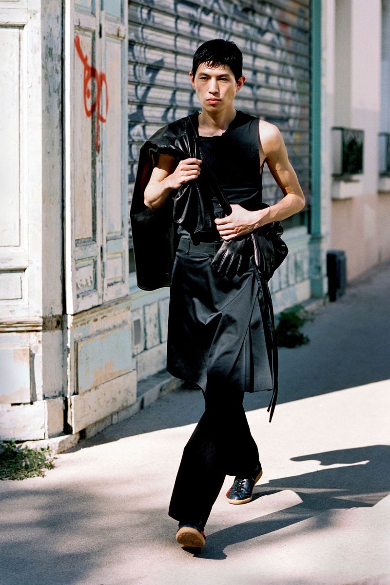 East Asian male model walking down a city sidewalk wearing an all-black sleeveless top, draped skirt trousers, and a leather jacket.