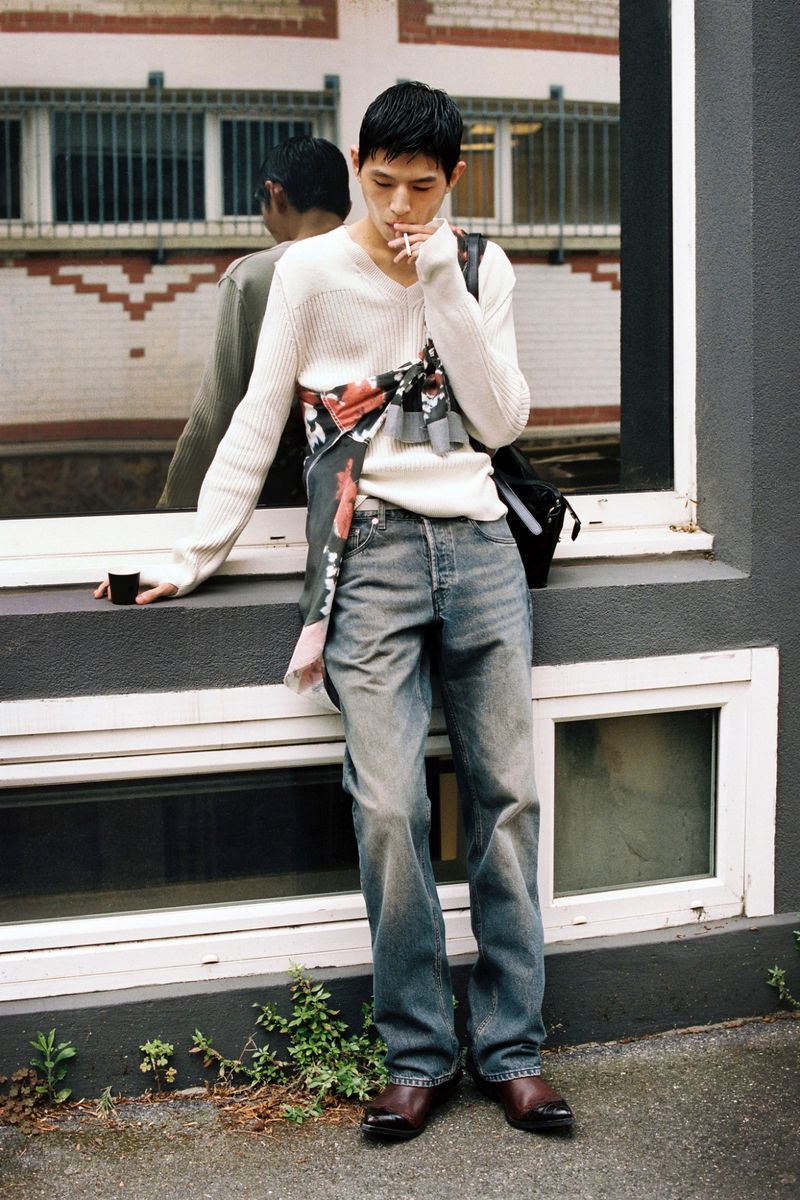 Man in a cream V-neck knit sweater and straight-leg blue jeans leaning against a dark exterior window frame while smoking.