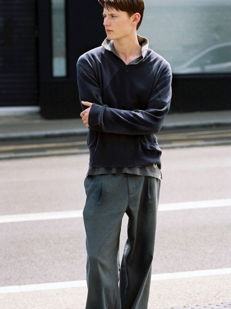 A man standing on a street wearing a dark v-neck sweater and grey trousers, with his arms crossed.