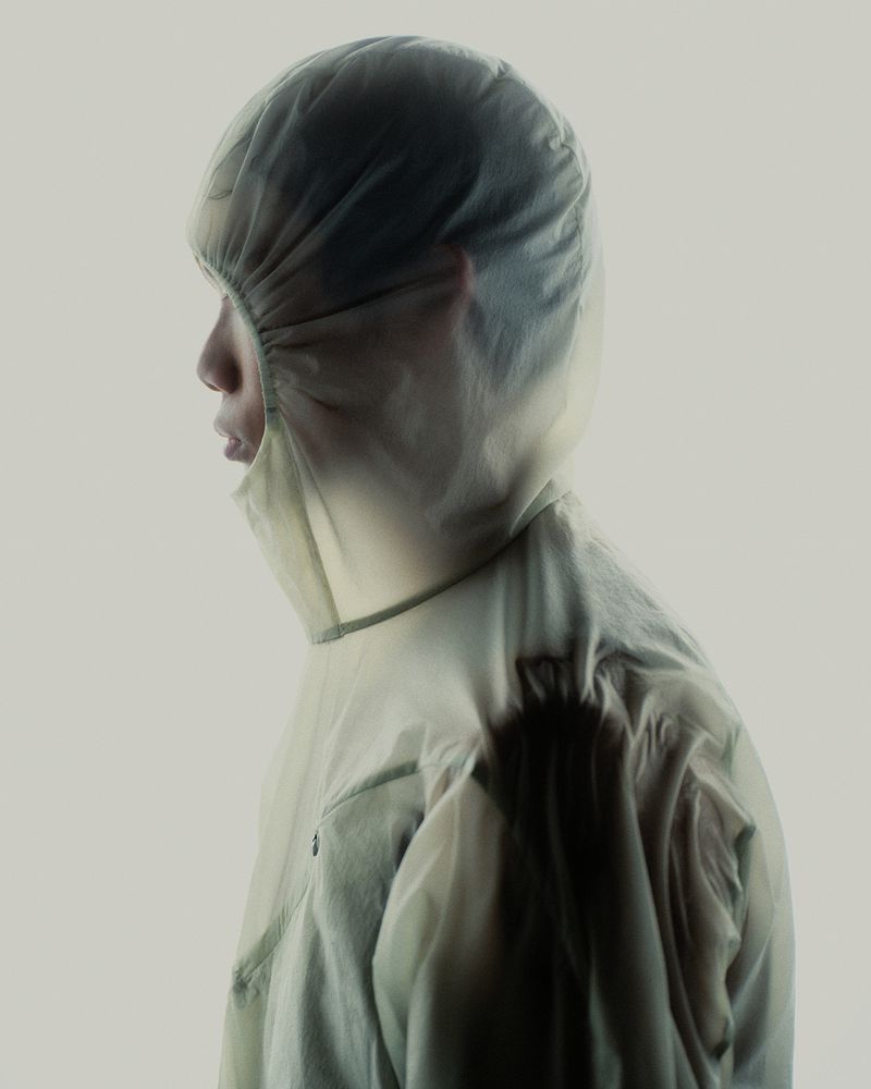 Profile of an East Asian man wearing a translucent pale green hooded jacket against a neutral studio backdrop.