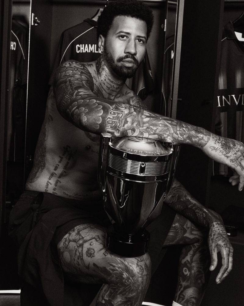 Miles Chamley-Watson sitting shirtless with extensive tattoos, resting his arm on an oversized trophy-shaped perfume bottle.