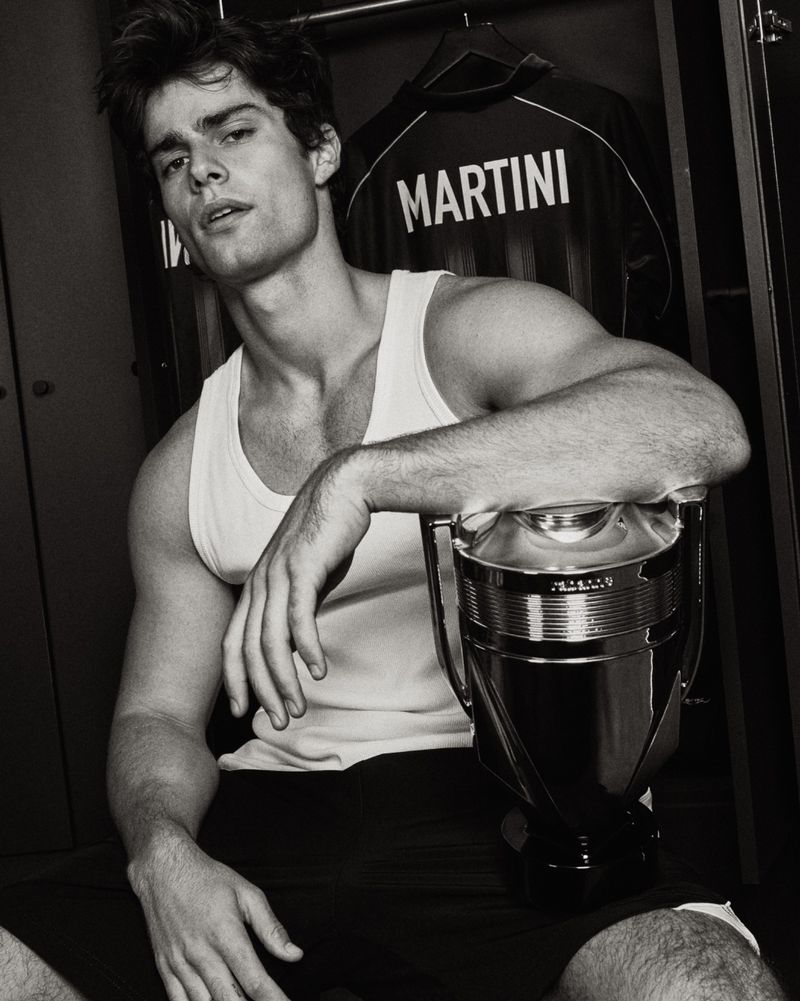 Corrado Martini in a white tank top leaning on a large silver Invictus trophy bottle inside an open locker room.