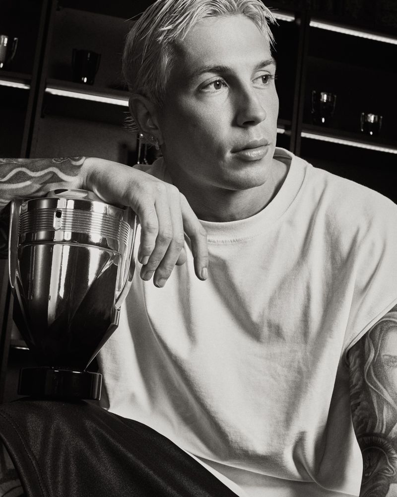Monochrome portrait of a blonde tattooed model in a white t-shirt resting his arm on a Rabanne Invictus trophy fragrance bottle.