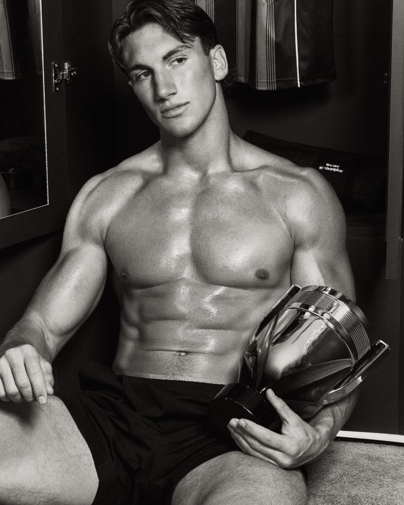 Shirtless muscular man seated in an open locker holding a polished metallic trophy bottle in a black and white portrait.