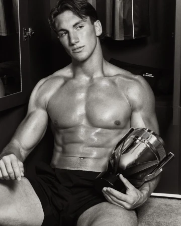 Shirtless muscular man seated in an open locker holding a polished metallic trophy bottle in a black and white portrait.