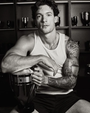 Monochrome portrait of a tattooed man in a white tank top resting his arm on a trophy in front of trophy-lined shelves.