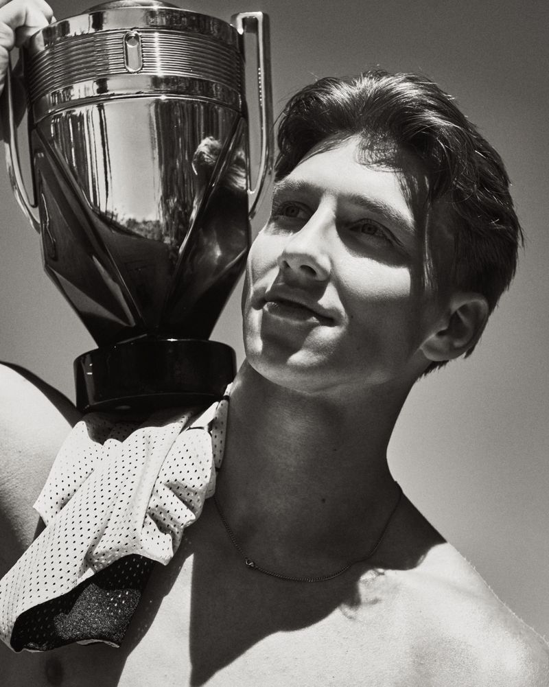 Black and white portrait of a shirtless young man balancing a large metallic trophy bottle on his shoulder in direct sunlight.