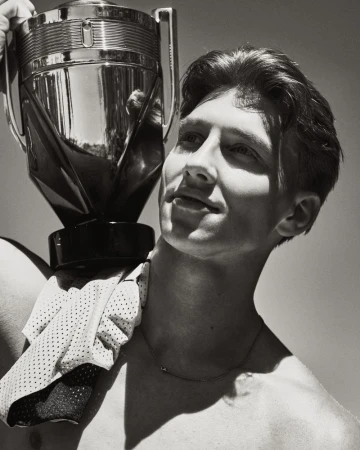Black and white portrait of a shirtless young man balancing a large metallic trophy bottle on his shoulder in direct sunlight.