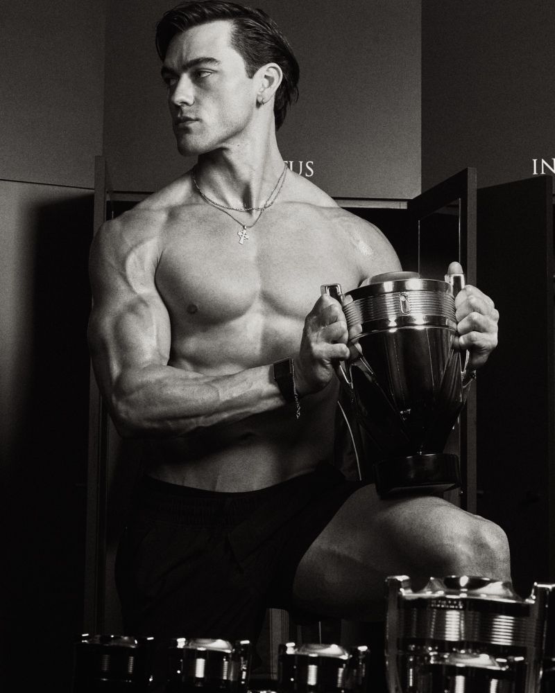 A shirtless muscular man holding a large trophy-shaped fragrance bottle in a high-contrast black-and-white studio shot.