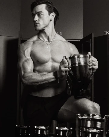 A shirtless muscular man holding a large trophy-shaped fragrance bottle in a high-contrast black-and-white studio shot.