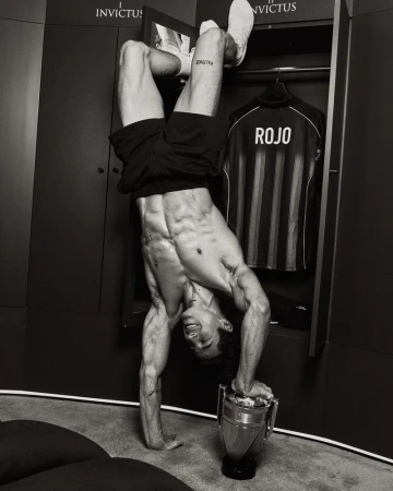 Shirtless athletic man balancing upside down in a handstand on a trophy inside a locker room setting.