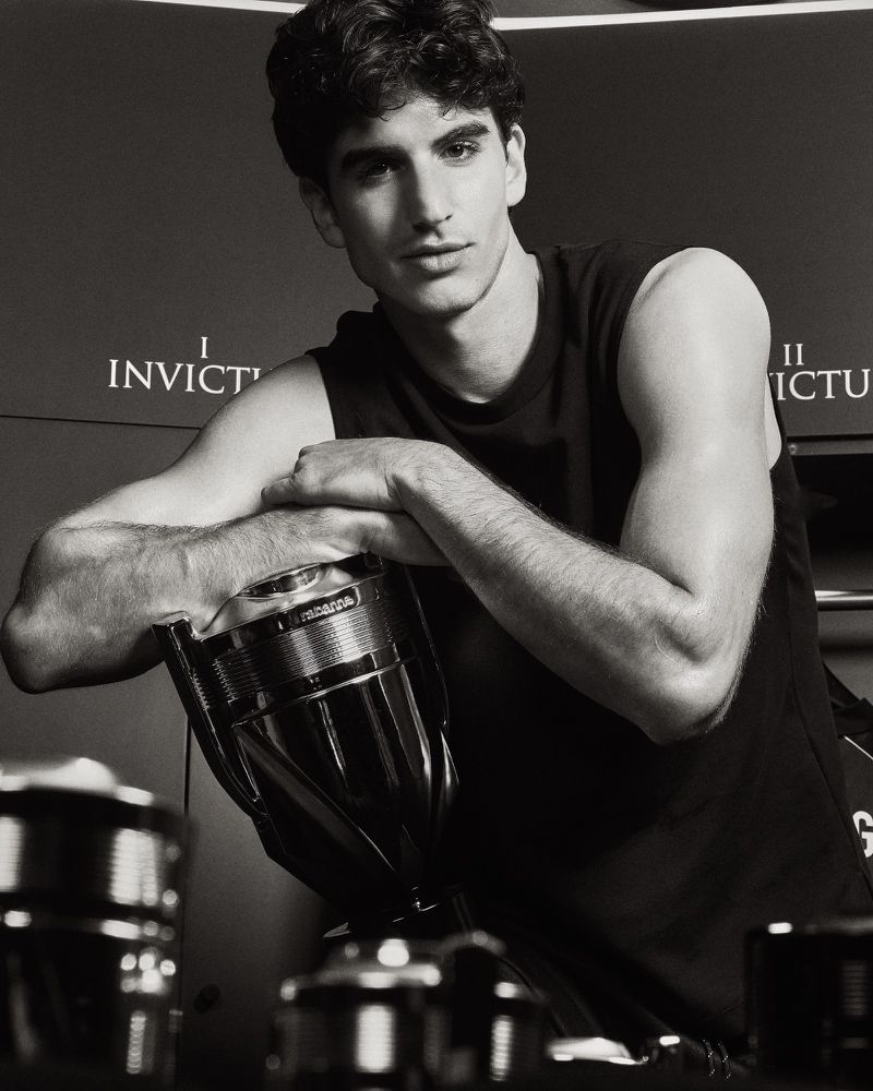 A man in a black sleeveless shirt leaning over an oversized Rabanne Invictus fragrance bottle in a black and white portrait.