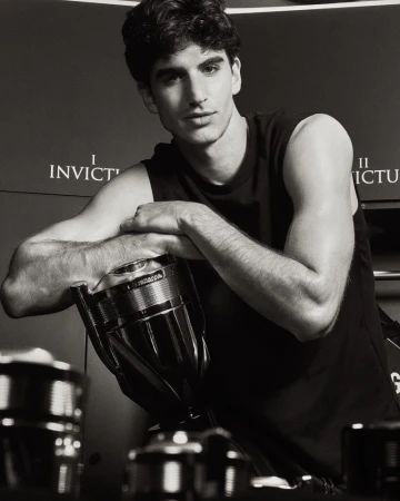 A man in a black sleeveless shirt leaning over an oversized Rabanne Invictus fragrance bottle in a black and white portrait.