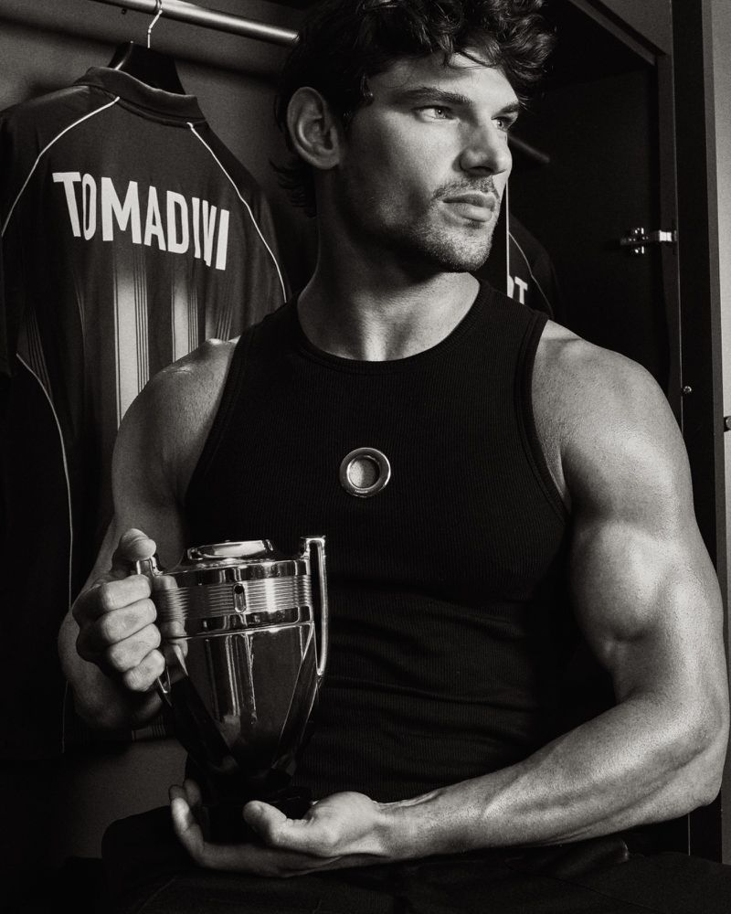 Monochrome portrait of model Stefano Tomadini holding a trophy-shaped fragrance bottle in a black tank top in a locker room.
