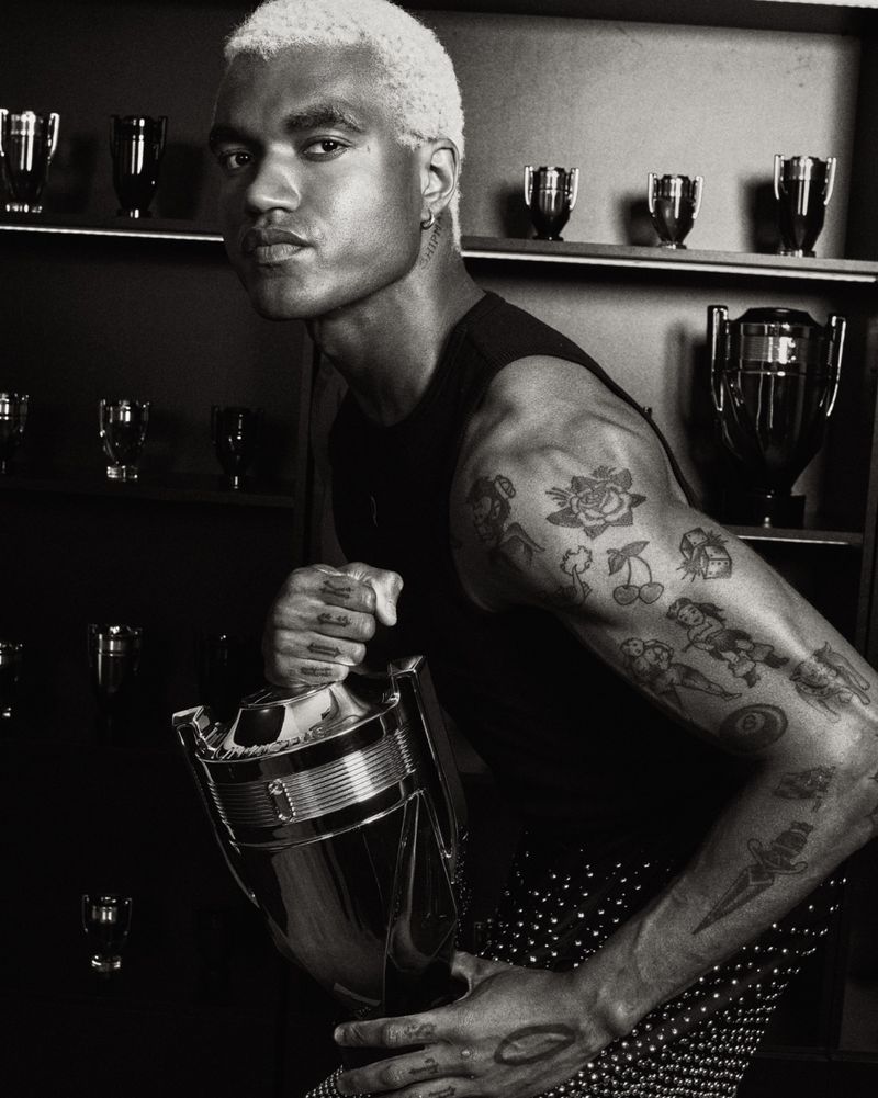 Miles Chamley-Watson with platinum hair and arm tattoos holding a trophy-shaped Rabanne Invictus bottle in monochrome.