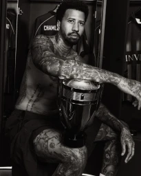 Miles Chamley-Watson sitting shirtless with extensive tattoos, resting his arm on an oversized trophy-shaped perfume bottle.