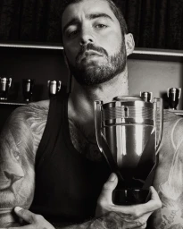 Tattooed man in a black ribbed tank top holding a metallic trophy-shaped Rabanne Invictus perfume bottle in black and white.
