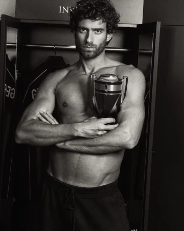 Shirtless man with curly hair and beard holding a trophy-shaped Rabanne Invictus fragrance bottle in black and white.