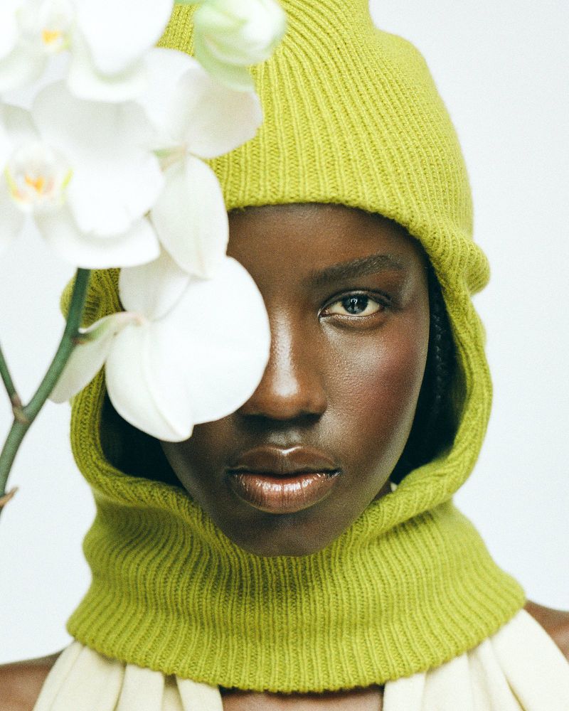 Close-up of Black woman in green balaclava, partially obscured by white orchid, against a white backdrop in a studio setting.