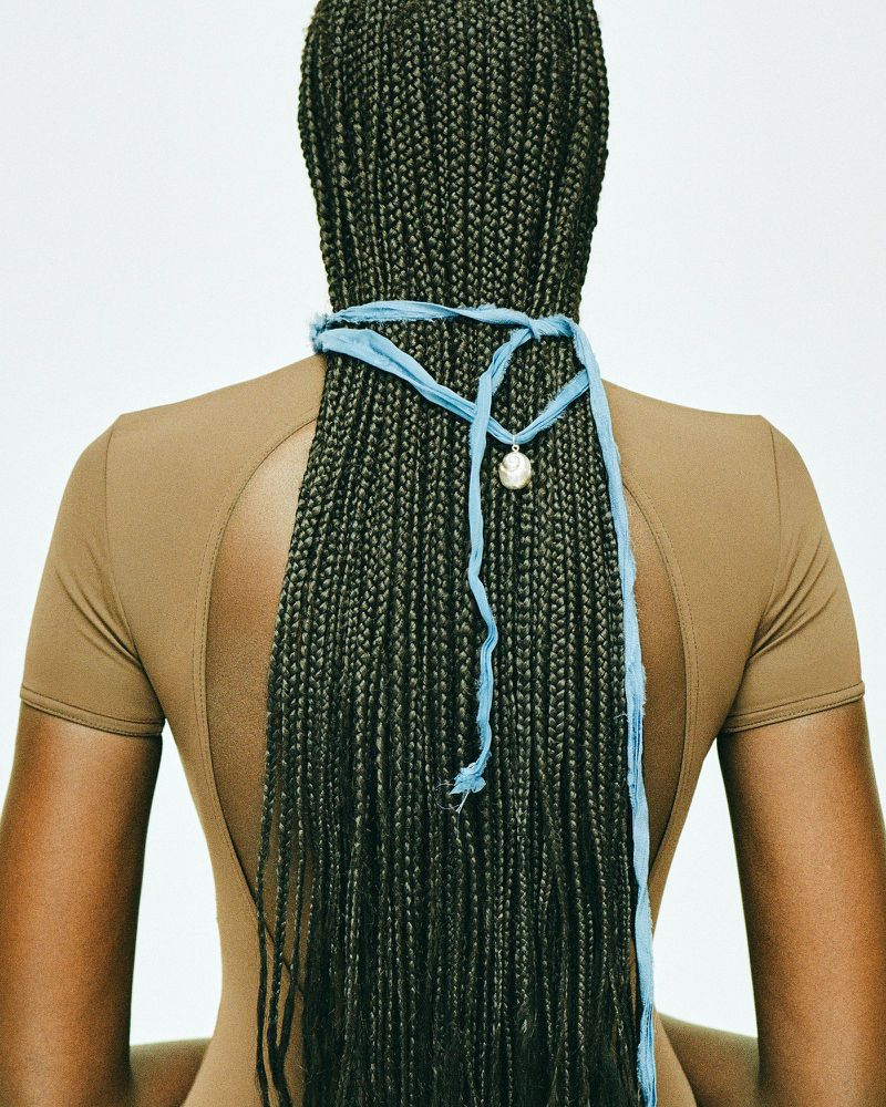 Model Mayor Dutie viewed from behind with long dark braids tied with a blue fabric ribbon and pearl pendant, wearing a tan open-back top.