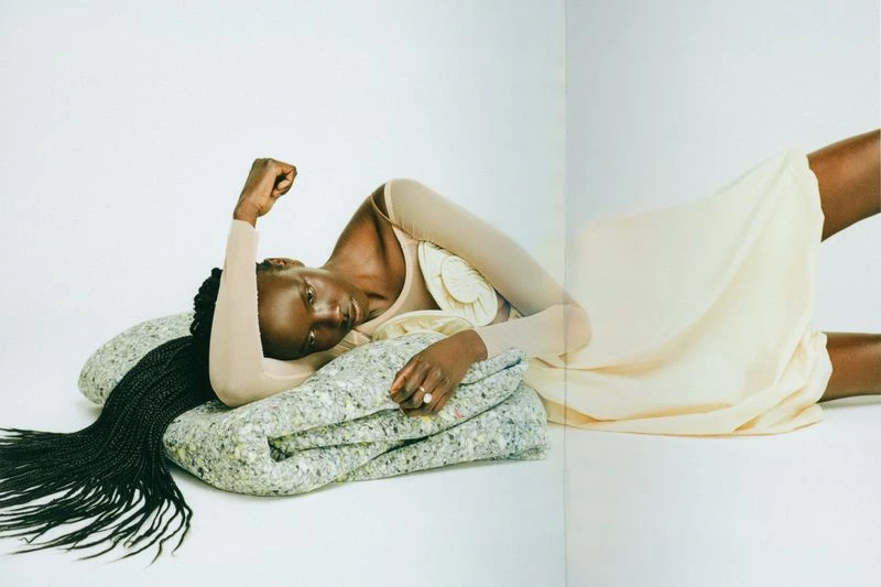 A Black woman lies on her side on a gray cushion wearing a beige top with a gathered white decoration and a pale yellow dress.