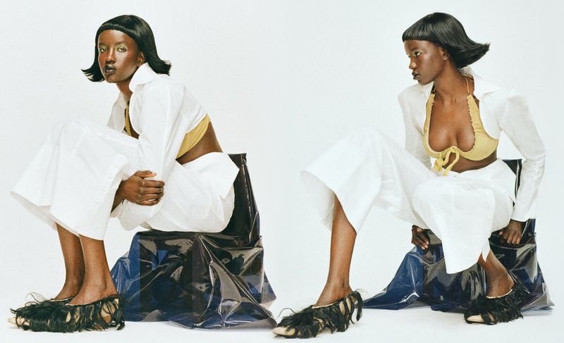 Two dark-skinned female models in white, wide-legged pants, open white shirts, gold bras, and black fringe shoes pose in a studio.