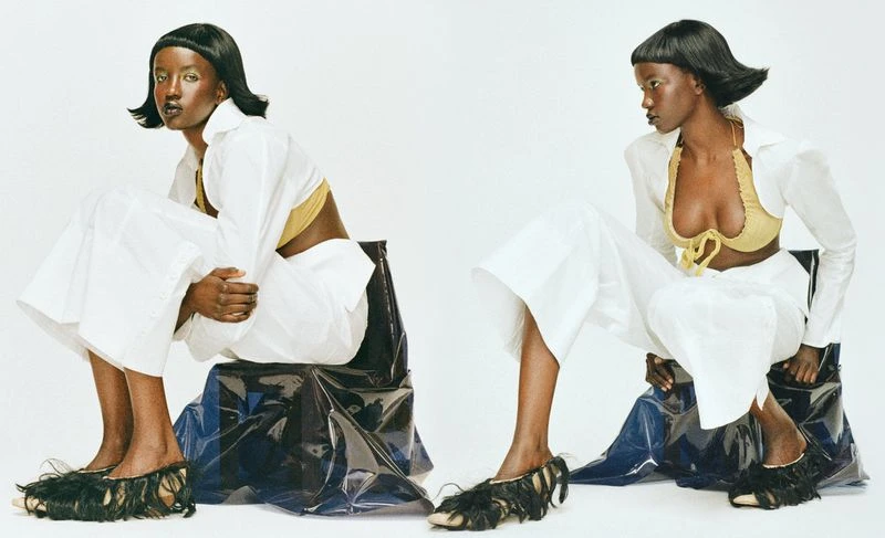 Two dark-skinned female models in white, wide-legged pants, open white shirts, gold bras, and black fringe shoes pose in a studio.