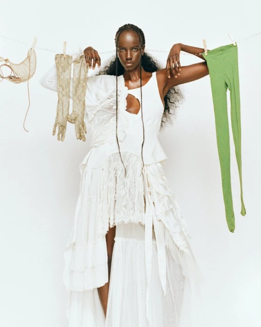 Fashion shot of a Black female model in white dress, posing with beige gloves and green leggings hanging from a clothesline on white.