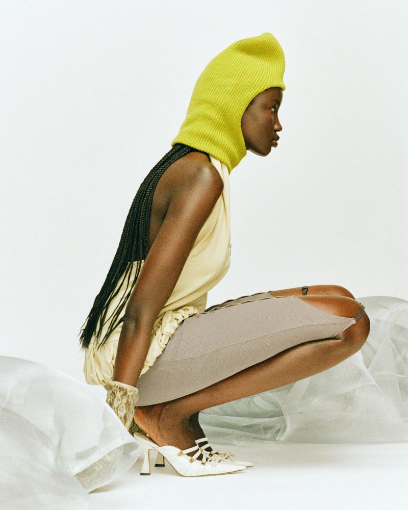 A dark-skinned woman with braided hair squats wearing a chartreuse knit balaclava, cream dress, grey skirt, and white heels in a photo studio.