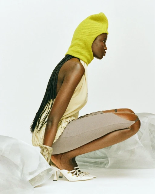 A dark-skinned woman with braided hair squats wearing a chartreuse knit balaclava, cream dress, grey skirt, and white heels in a photo studio.