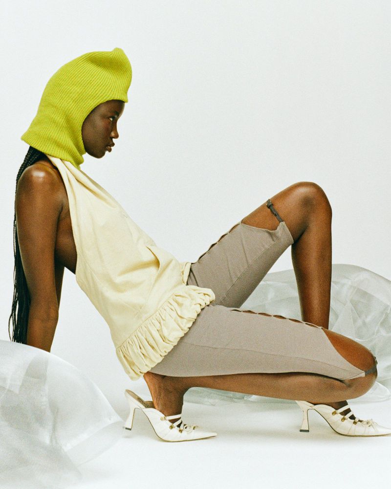 Woman poses in a light yellow top, grey shorts with side slits, and white heels, accessorized with a chartreuse knit hood in a studio.