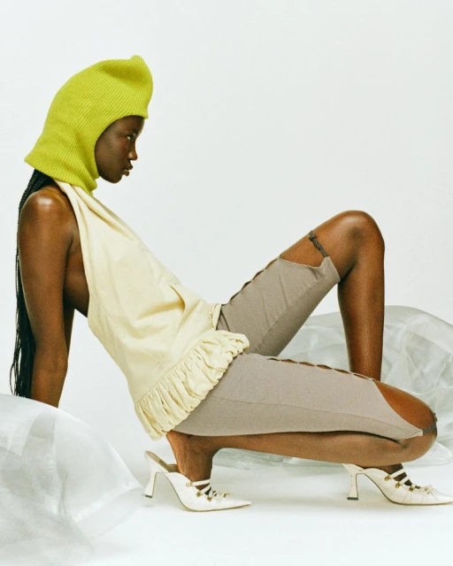 Woman poses in a light yellow top, grey shorts with side slits, and white heels, accessorized with a chartreuse knit hood in a studio.