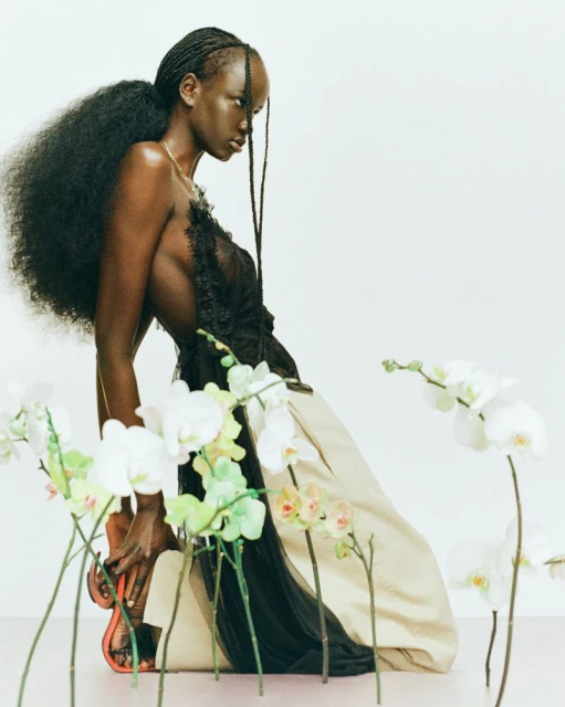 Woman in an elegant black top and tan skirt kneeling near orchids, showcased in a brightly lit studio with a seamless backdrop.