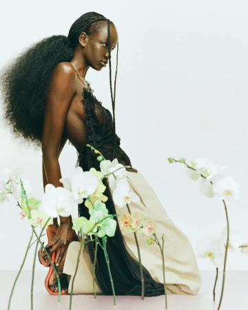 Woman in an elegant black top and tan skirt kneeling near orchids, showcased in a brightly lit studio with a seamless backdrop.