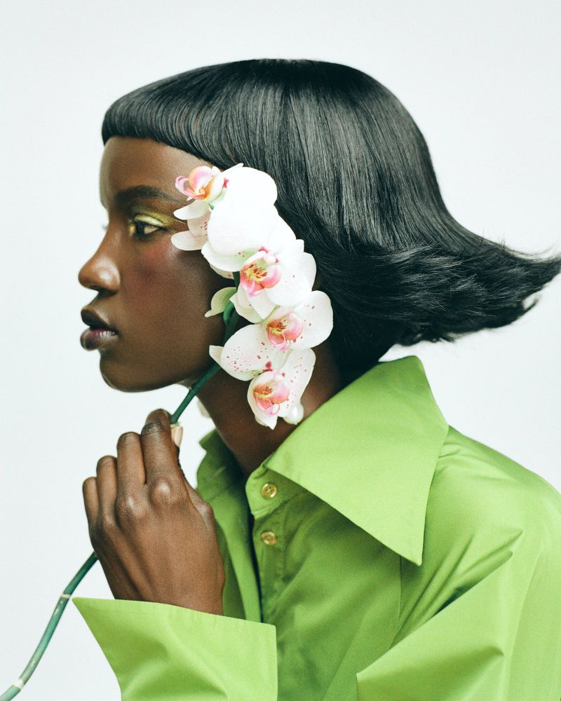Elegant portrait of a dark-skinned woman in a green shirt, adorned with orchids in her hair, showcasing a beauty and fashion aesthetic.