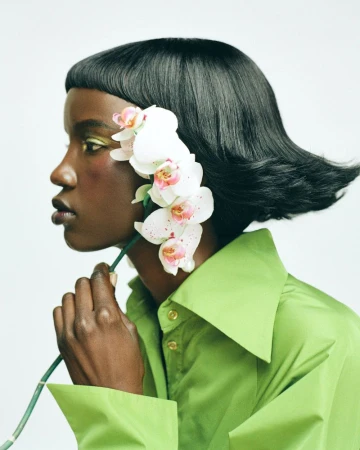 Elegant portrait of a dark-skinned woman in a green shirt, adorned with orchids in her hair, showcasing a beauty and fashion aesthetic.