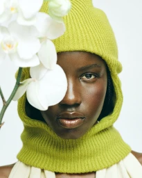 Close-up portrait of a Black woman wearing a lime green knit balaclava partially obscured by white orchid flowers