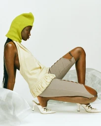Model wearing a chartreuse balaclava and cream halter top crouches on a white studio background.