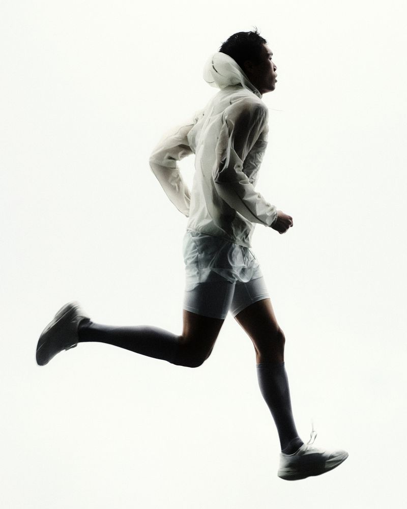 Runner captured mid-stride wearing a translucent hooded white jacket, white shorts, dark socks, and white sneakers on a white background.