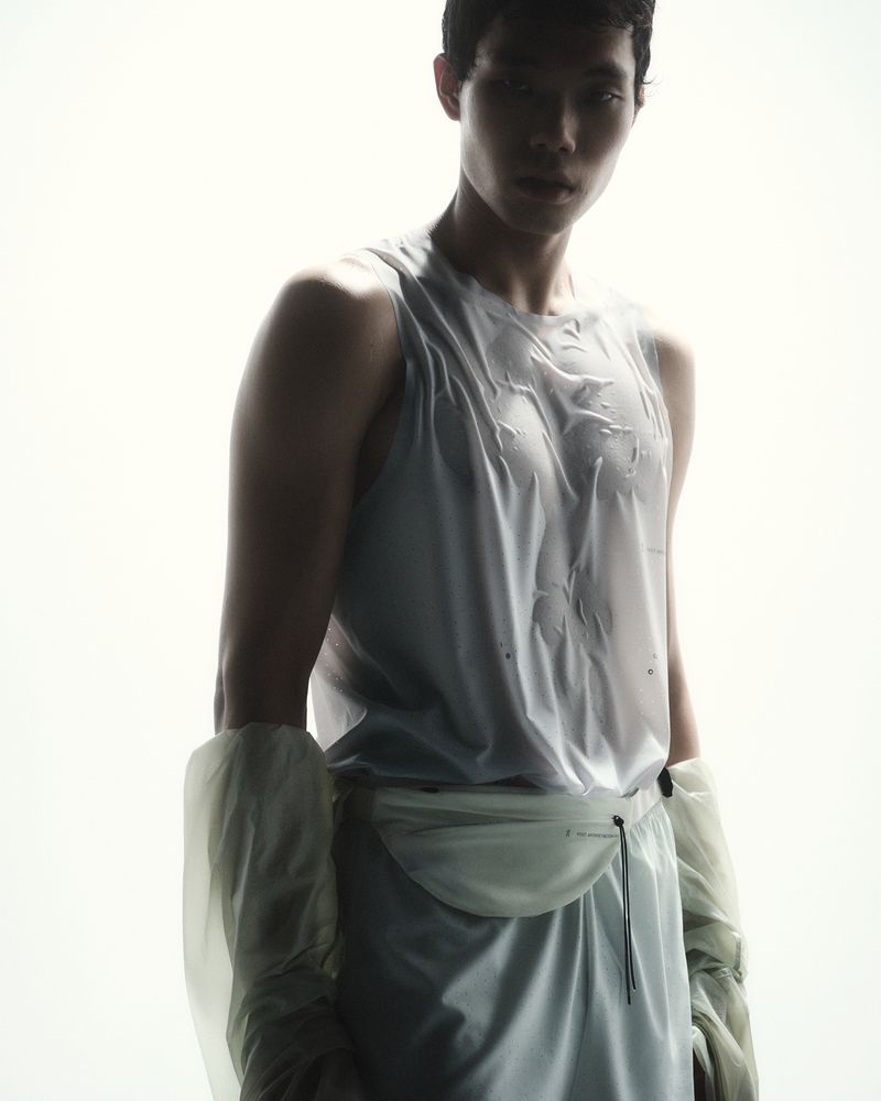 East Asian male model wearing a sheer white tank top and technical waist pack against a bright white backlit background.