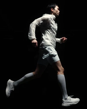 Male runner mid-stride wearing a white technical jacket, white shorts, tall socks, and running sneakers against a black background.