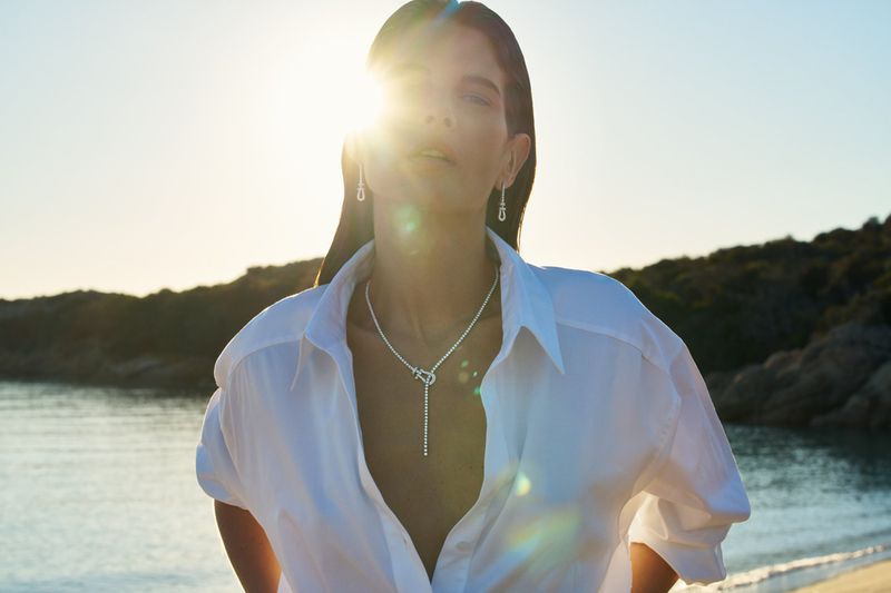 Model Monica Cima wearing an unbuttoned white shirt and diamond jewelry against a sunlit coastal background.