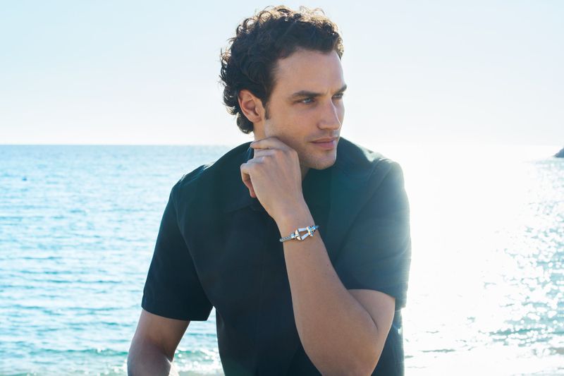 Close-up portrait of a man wearing a dark short-sleeve shirt and a silver bracelet outdoors by the ocean.