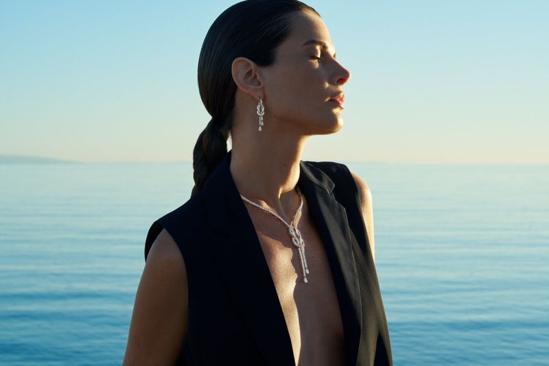 Side profile of a woman with slicked hair wearing a black sleeveless blazer, diamond necklace, and earrings against open blue sea.