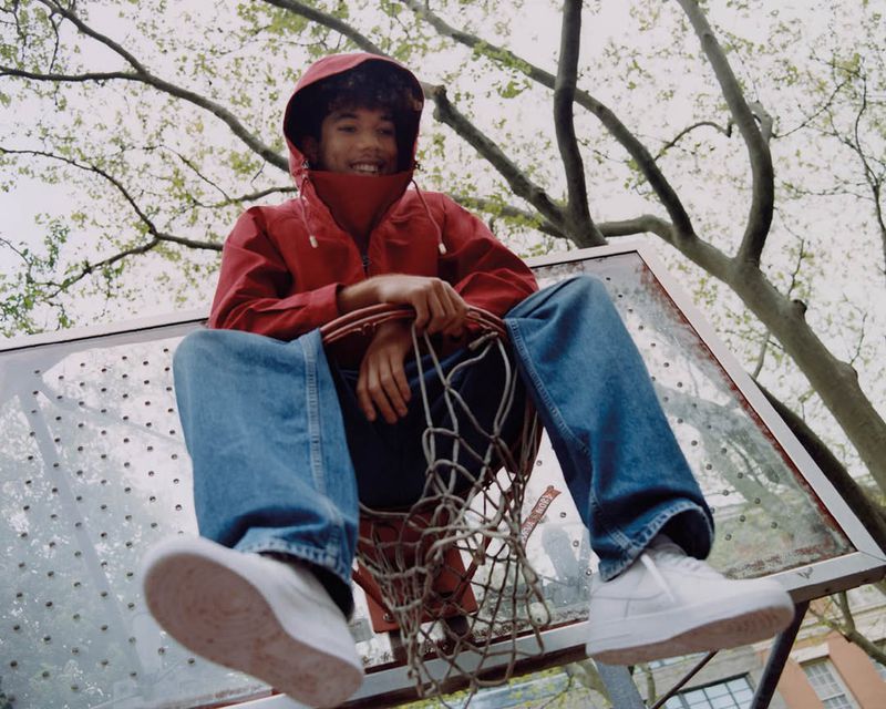 Young man in a red hooded jacket sitting on a basketball hoop outdoors, wearing jeans and white sneakers, smiling at the camera.