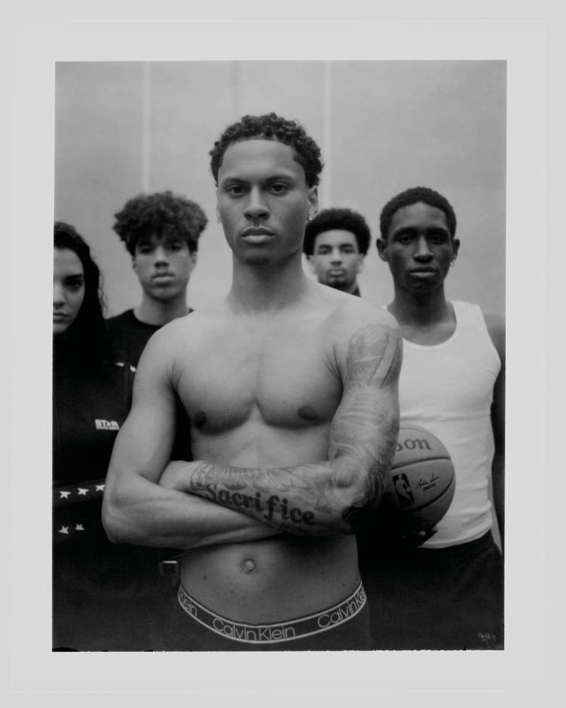 Black and white portrait of shirtless athlete posing in front of team with basketball, showcasing toned physique and ‘Sacrifice’ tattoo.